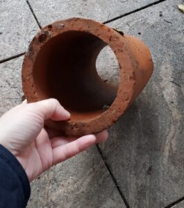 A white hand holds an orange-ish clay pipe against a backdrop of grey patio tile.